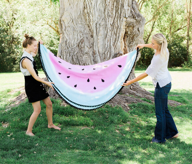 2 women holding up a watermelon designed picnic blanket by a tree.